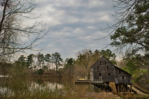 A cloudy landscape at the tranquil pond of historic Yates Mill Park framed between trees during Spring with the rebuilt timber gristmill in Raleigh, North Carolina.