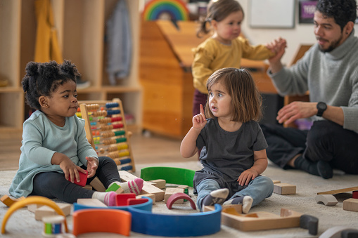 Daycare children are seen sitting on the floor as they pay with wooden blocks together.  The toddlers are each dressed casually and are focused on their play as they interreact with one another.  Their male educator can be seen in the background playing with other children.