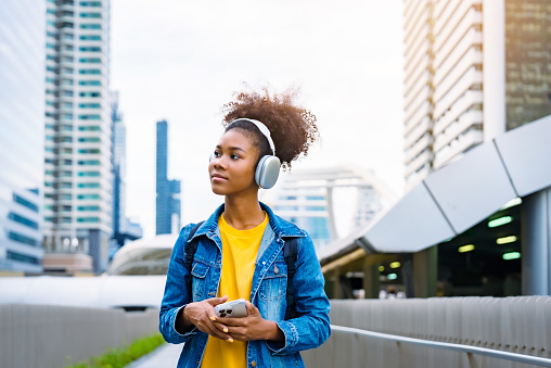 Cheerful black student girl wearing headset and using mobile smartphone, Walking at college building outdoor, Happy and smiling woman teenager. Modern education