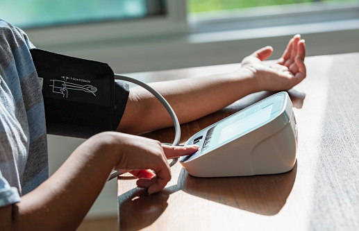 The arms of a young woman measuring her blood pressure at home. She is sitting at a table with the blood pressure cuff on her arm. Her forearm and the digital display are on the table. With the other hand, she is pressing the start button on the device.