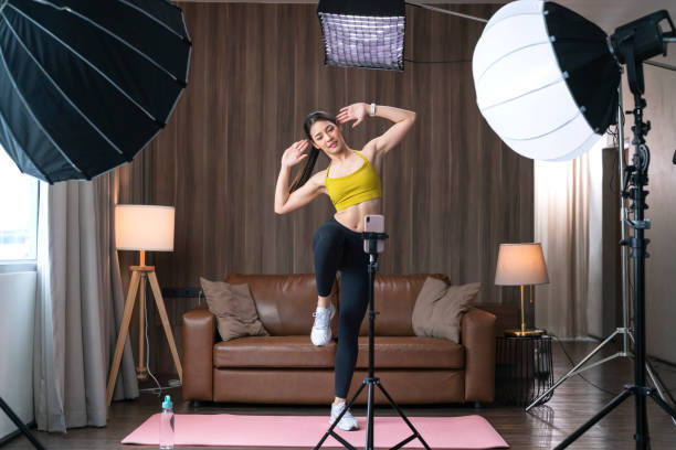 Cheerful woman wearing sports top and leggings standing active sport posture in living room and recording video using smartphone on tripod with studio lighting. Fitness blogger streaming online from home conversación de ropa deportiva de mujer activa de asia hablando con la cámara y explicando cómo trabajar para su estudiante seguidor en línea. influencer de deporte saludable en la sala de estar de casa. concepto de clase de culturismo en línea de ent