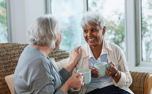 Two multiracial senior women sitting together on a sofa conversing over coffee. The African-American woman is in her 70s. The other woman, in her 60s, has her hand on her friend's shoulder, and they are smiling.