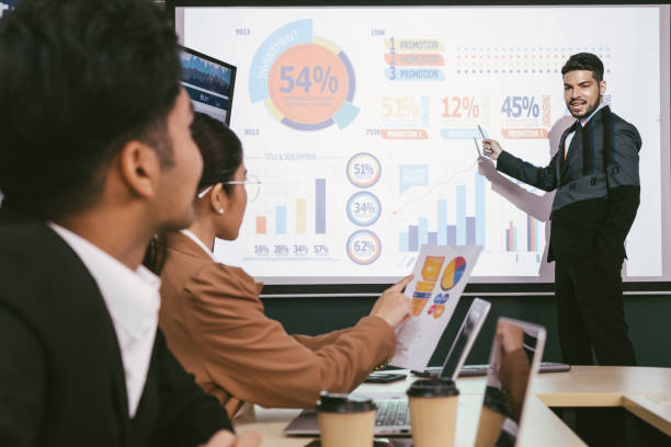 istock A cheerful and confident Asian businessman stands, presenting bar charts data from the projector screen to his office colleagues. Asian businessman leader role at the meeting. 1472027609