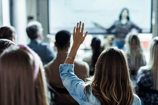 Rear view of a female freelancer raising her hands to ask a question on a seminar in board room.