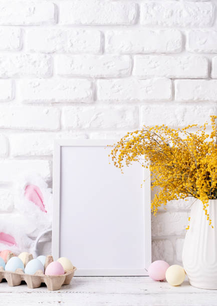 istock Pastel colored Easter eggs and mimosa flowers with blank white frame for mockup design, front view on white brick wall background 1472034421