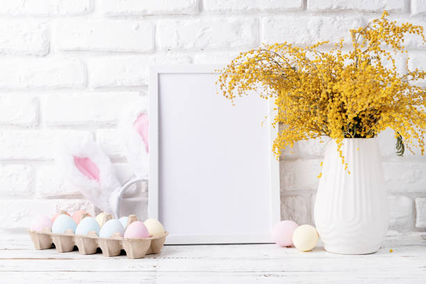 istock Pastel colored Easter eggs and mimosa flowers with blank white frame for mockup design, front view on white brick wall background 1472036238