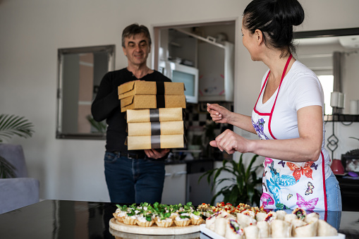 A young woman who is preparing catering food is completing new orders by packing them in paper boxes delivered by a male colleague.