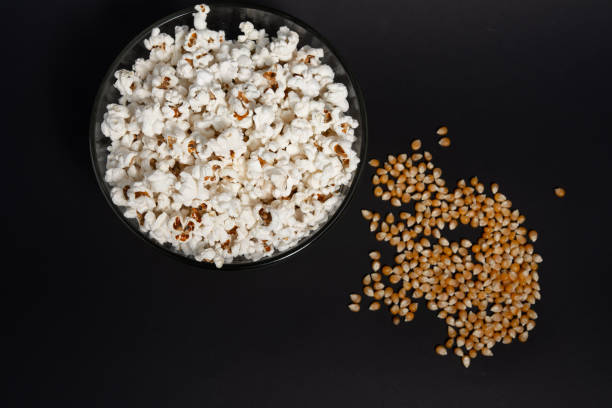 pipoca pronta para comer em uma tigela de vidro e grãos de milho em um fundo preto. - popcorn black dietary fiber refreshment - fotografias e filmes do acervo