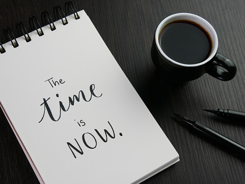 THE TIME IS NOW. black lettering in notepad with cup of espresso and pen on black wooden desk
