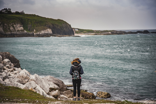 A tourist or hicker takes a break while hiking on a wild trail to admire the dramatic cliffs and gloomy Irish landscape by the the Giant's Causeway. A pensive woman staring at breathtaking ocean is meditating about life  - Bushmills, Northern Ireland, United Kingdom