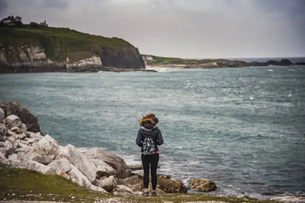 Photo of A tourist or hicker takes a break while hiking on a wild trail to admire the dramatic cliffs and gloomy Irish landscape by the the Giant's Causeway. A pensive woman staring at breathtaking ocean is meditating about life  - Bushmills, Northern Ireland