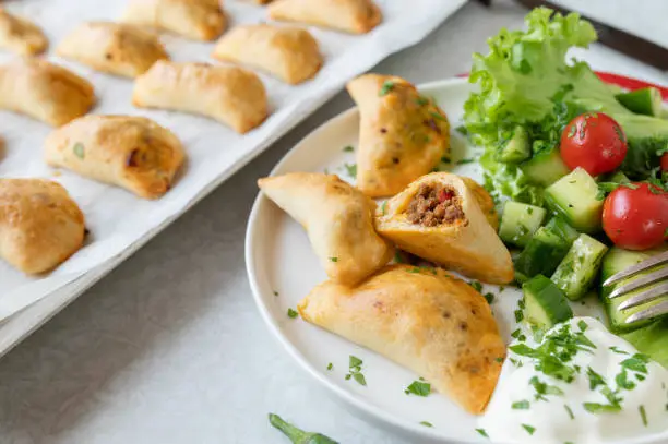 Delicious homemade empanadas with ground beef, vegetable, cheese filling. Served with a fresh side salad on a white plate with sheet pan in the background on light table background. Top view