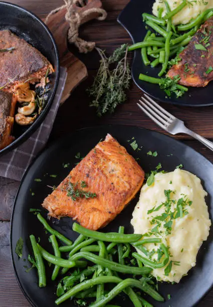 Delicious salmon dish, fried with skin and served with fresh mashed potatoes and green beans on a dark plate on rustic and wooden table background with frying pan. Top view and closeup