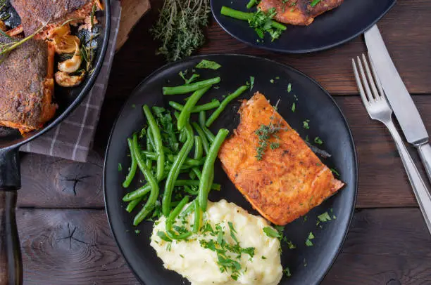 Delicious and gluten free fish dish with  pan fried salmon fillet with green beans and mashed potatoes. Served on a dark plate on rustic and wooden background. Table top view
