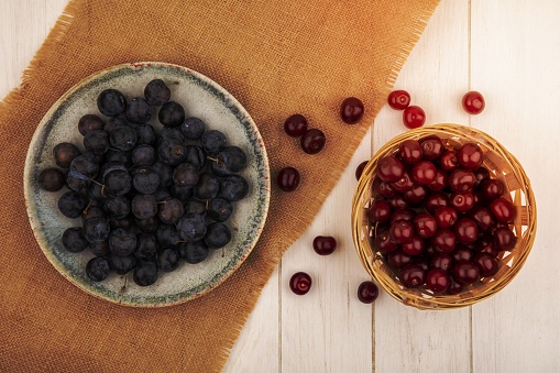 top view of the small sour blue-black fruit sloes on a bowl on a sack cloth with red cherries on a bucket on a white background