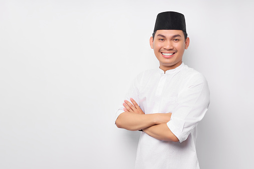 Optimistic handsome young Asian Muslim man in Arabic costume standing with crossed arms and looking at camera with toothy smile isolated on white background. People religious Islamic lifestyle concept