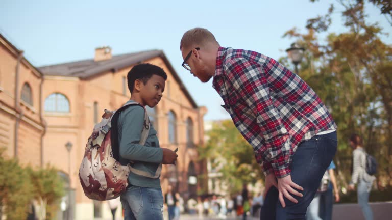 Young strict male teacher yelling at boy while standing near school. Realtime