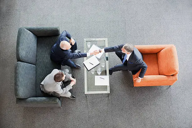 Photo of Businessmen shaking hands across coffee table in office lobby