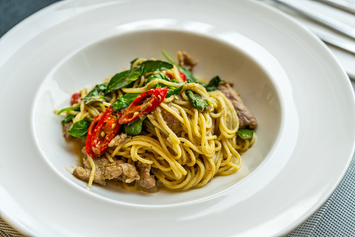 Delicious Thai fusion food, stirred fried spaghetti with thai green curry and meat. Beautiful Thai food in modern white dish on a table in restaurant, natural light from window.