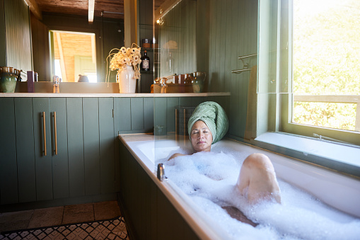 Mature woman with her hair wrapped in a towel lying with her eyes closed while relaxing in her bath at home