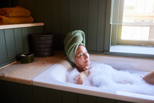 Mature woman with her hair wrapped in a towel lying with her eyes closed and soaking in her bathtub at home