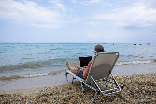 Mid-adult Caucasian man, working on laptop, while listening to music on wireless headphones, while sitting on the outdoor chair at the beach, during his vacation