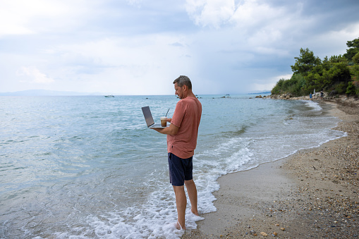 Mid-adult Caucasian man, working on a laptop, while drinking iced coffee on the beach