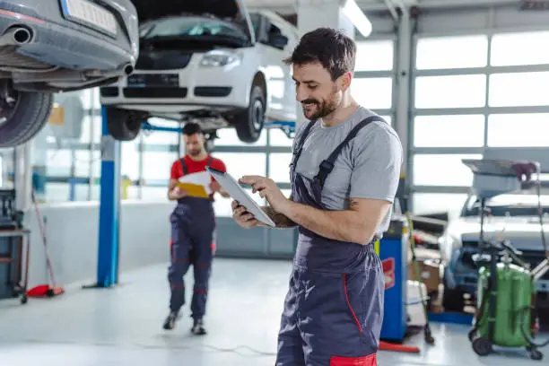 Smiling auto mechanic in auto service holding digital tablet. In background is the other worker