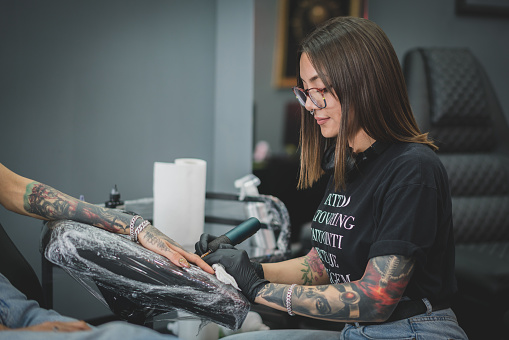 Caucasian female tattoo artist, tattooing the female customer in her tattoo studio