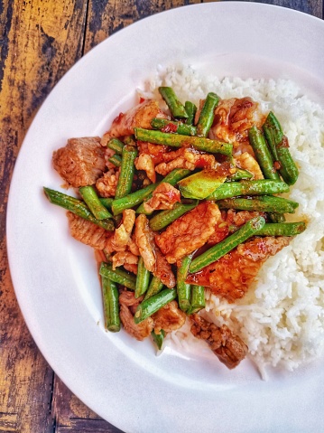 Stir-fried pork with red curry paste with yard long beans over rice in a white plate placed on a table on a wooden floor. Take photo in Bangkok Thailand
