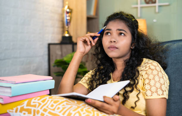 istock thoughtful girl while reading book at home while sitting on sofa during exam preparation - concept of hard working, exam preparation and education. 1472089781