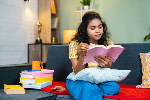 istock Serious young girl reading book for examination while sitting on sofa at home - concept of education, preparation and hobbies 1472091096