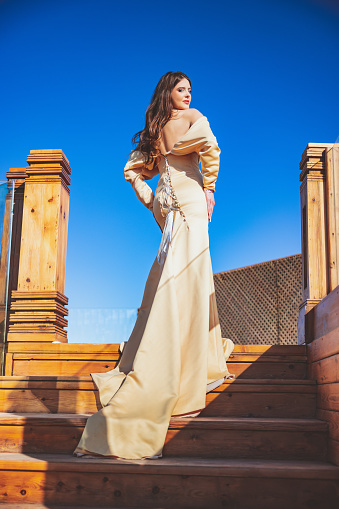 Rear view on beautiful young fashion model with brown hair down wearing beige backless dress of shoulder, standing on the top of the wooden stairs on a fine day and turning to look at camera over shoulder, beauty and fashion industry