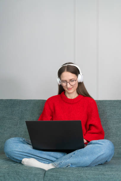 赤いセーター、ジーンズ、白いヘッドフォンを着た若い女性が、自宅のソファでネットサーフィンをしている。ホームオフィスで働く女の子。ラップトップとインターネットを使用するフリ� - yoga business women living room ストックフォトと画像
