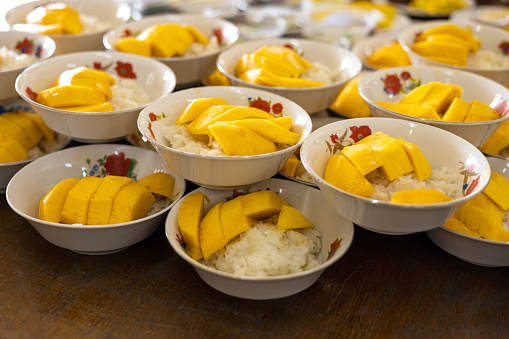 Ripe mango sticky rice snacks packed in many white bowls are stacked on wooden tables, ready to be served to people at feasts common in rural Thailand.