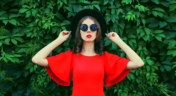 Portrait of beautiful young woman posing in red dress, black round hat on green leaves wall background