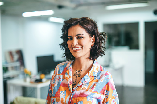 Cheerful businesswoman with tattoo, wearing colorful shirt, smiling at camera in office
