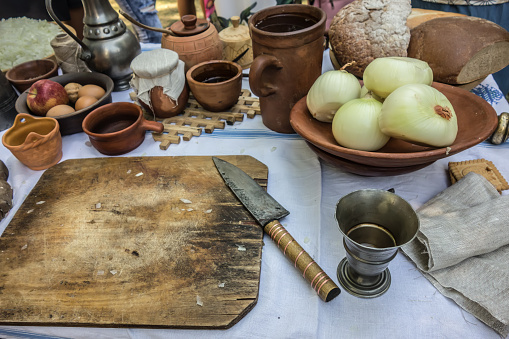 Rustic table during cooking. Close-up of an old wooden cutting board with a kitchen knife. Bread, onions, apples, clay and silver dishes are on the table.