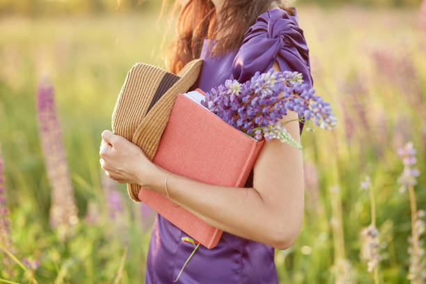 woman in summer sunset field with bouquet of wildflowers - lupines, hat and pink notebook. closeness to nature, self-discovery concept. relax and welll-being concept. sunny day, good mood concept - day to sunset imagens e fotografias de stock