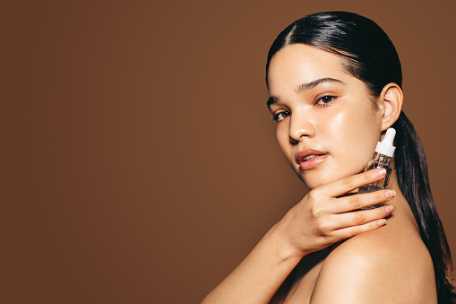 Using female cosmetic products for healthy skin: Portrait of a confident young woman holding face serum in a studio. Hispanic woman with beautiful skin showcasing her healthy skincare routine.