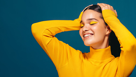 Woman in her 20’s smiling while standing in a studio with makeup on her face. This radiant young woman embraces her beauty unapologetically and expresses herself through vibrant colours.