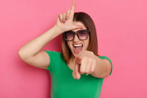 You are looser. Portrait of excited attractive young girl with brown hair wearing green casual t shirt and sunglasses, showing looser gesture and pointing at camera, posing against pink wall