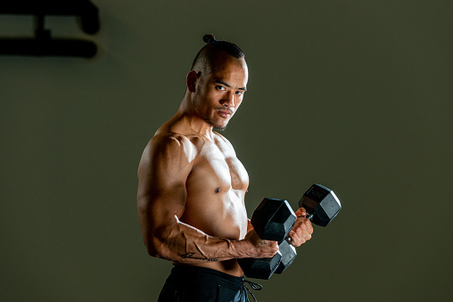 Muscular man working out in gym doing exercises with barbell, weights at biceps, looking at camera. Side view.