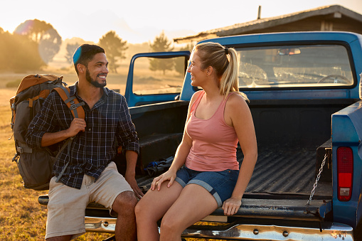 Couple Sitting On Tailgate Of Pick Up Truck With Rucksack On Road Trip To Cabin In Countryside