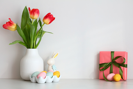 Vase of tulips, Easter bunny, colorful eggs, gift box on the table against the white wall.