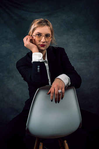 Fashion beauty portrait of a model in a classic suit white shirt posing on a chair. isolated on dark background. business woman. boss