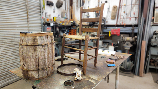 Active senior man restoring old nail keg and wooden back chairs with leather hide seat bottoms.  He has the keg and chairs on a worktable in his workshop.