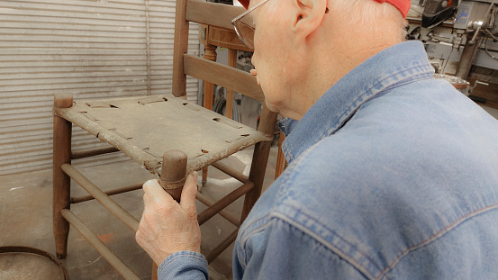 Active senior man restoring old nail keg and wooden back chairs with leather hide seat bottoms.  He has the keg and chairs on a worktable in his workshop.