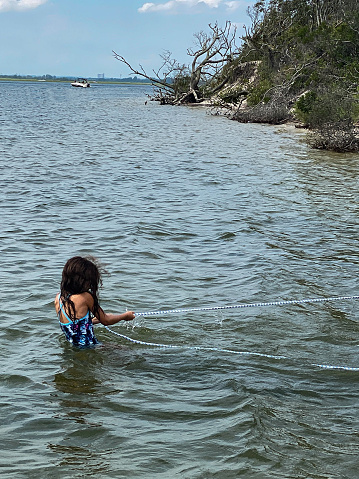 a young girl pulling something heavy out of frame while wading in the water with land and a boat in the background.