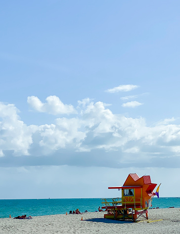 Fun photograph of a colorful lifeguard house on a sandy beach with bright blue waters, big fluffy clouds  hang in the clear blue sky.